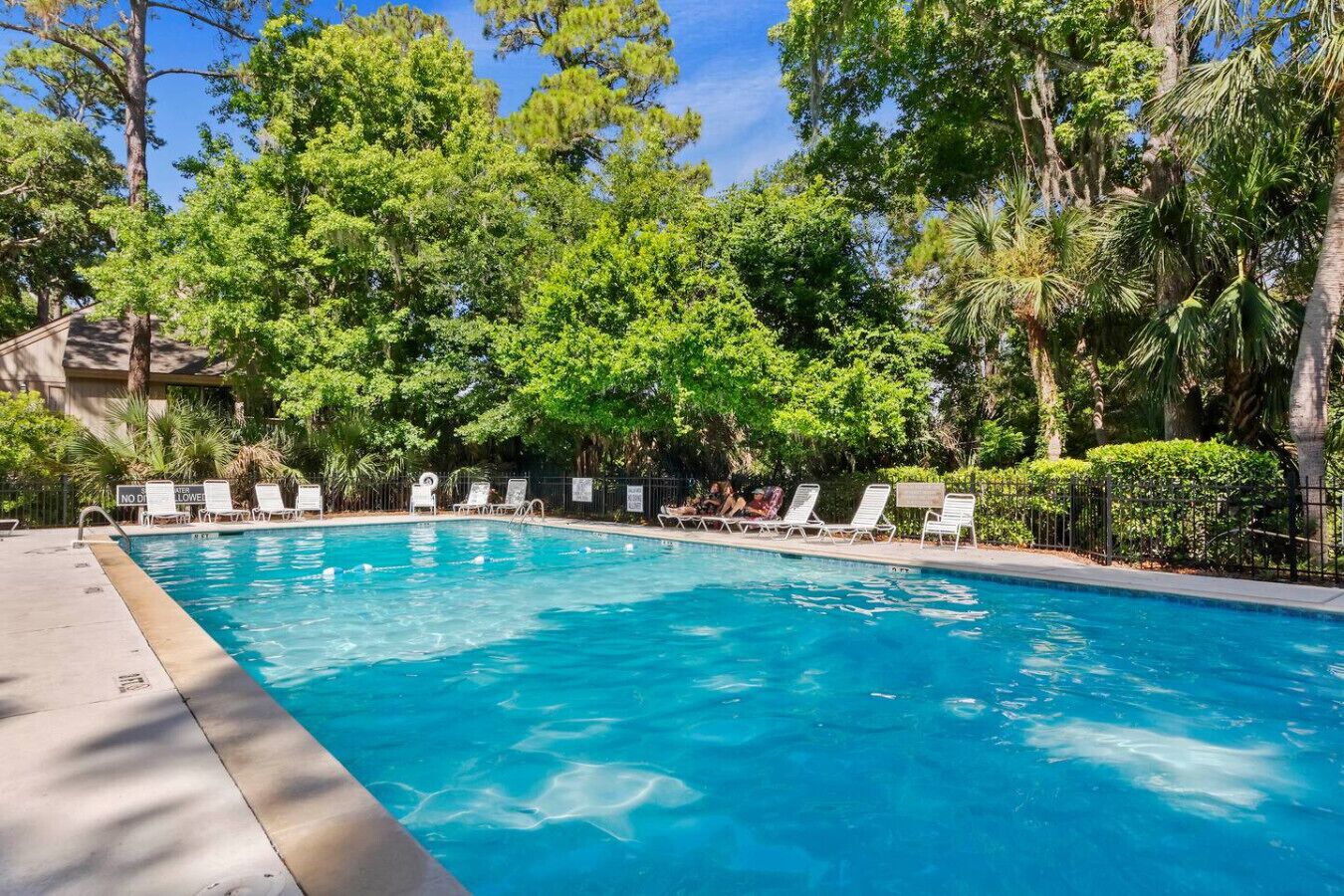 Piscina com espreguiçadeiras e cerca, rodeada por árvores.
