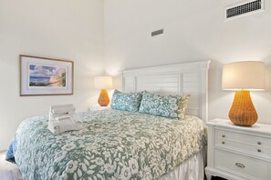 Una cama bien hecha con cubrecama floral, dos lámparas de noche, un cuadro en la pared y una mesita de noche blanca con cajones.