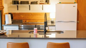 A modern kitchen with a central island, two bar stools, a refrigerator, and shelves with kitchen items. - Cute Little Cabin (Centerville)