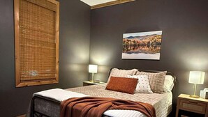 A bedroom with a bed, a ceiling fan, a window with blinds, a nightstand, and a wall-mounted lamp. - Cute Little Cabin (Centerville)