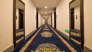 Corredor de hotel comprido e estreito com carpete estampado, paredes com espelhos e uma série de portas em ambos os lados.