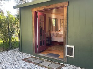 A cozy, single-room cabin with a wooden door leading to a bed, a small table, and a window with curtains.