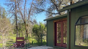 Una cabaña rústica con un sendero de grava que conduce a una puerta roja, rodeada de árboles y un jardín.