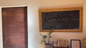 Un letrero de bienvenida sobre una puerta de madera, una mesita con una planta, un libro enmarcado y un cartel con normas.