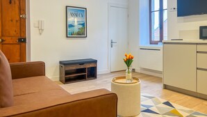 A modern living room with a brown sofa, a small wooden bench, a round coffee table with a vase of flowers, a framed picture on the wall, and a kitchen area with a TV and cabinets. - Ushuaia calme rue Carnot (Annecy)