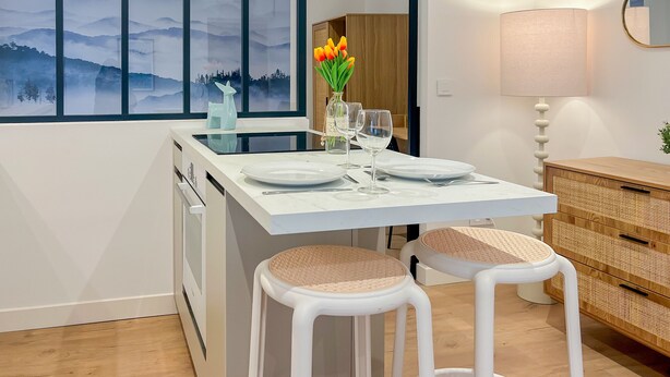 A modern kitchen with a dining area, featuring a large window with a scenic view, a white dining table set with plates and glasses, and a wooden cabinet with a woven basket. - Ushuaia calme rue Carnot (Annecy)