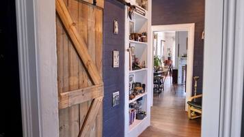 A hallway with a wooden door, shelves with various items, and a dining area in the background.