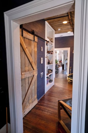Un couloir avec une porte en bois, des étagères remplies de divers objets et, au fond, un coin repas.
