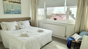 Un dormitorio ordenado con una cama grande, una ventana con vistas a edificios y árboles, y un cuadro en la pared.