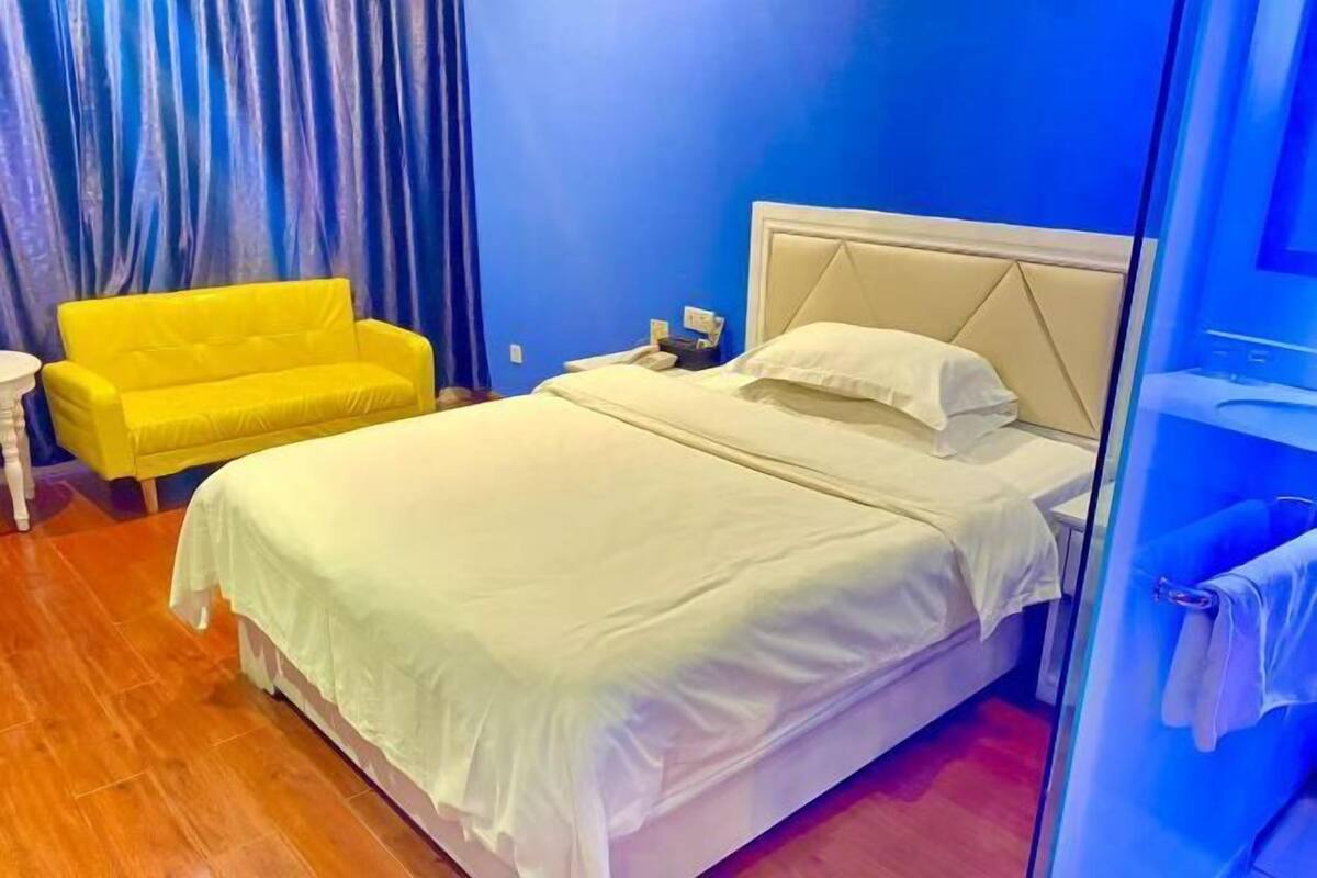 A hotel room with a bed, a yellow armchair, a small table, and a glass partition.