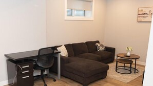Ein modernes Wohnzimmer mit einem braunen Ecksofa, einem schwarzen Schreibtisch mit Stuhl, einem kleinen runden Tisch mit einer Vase und einem Fenster mit Jalousien.