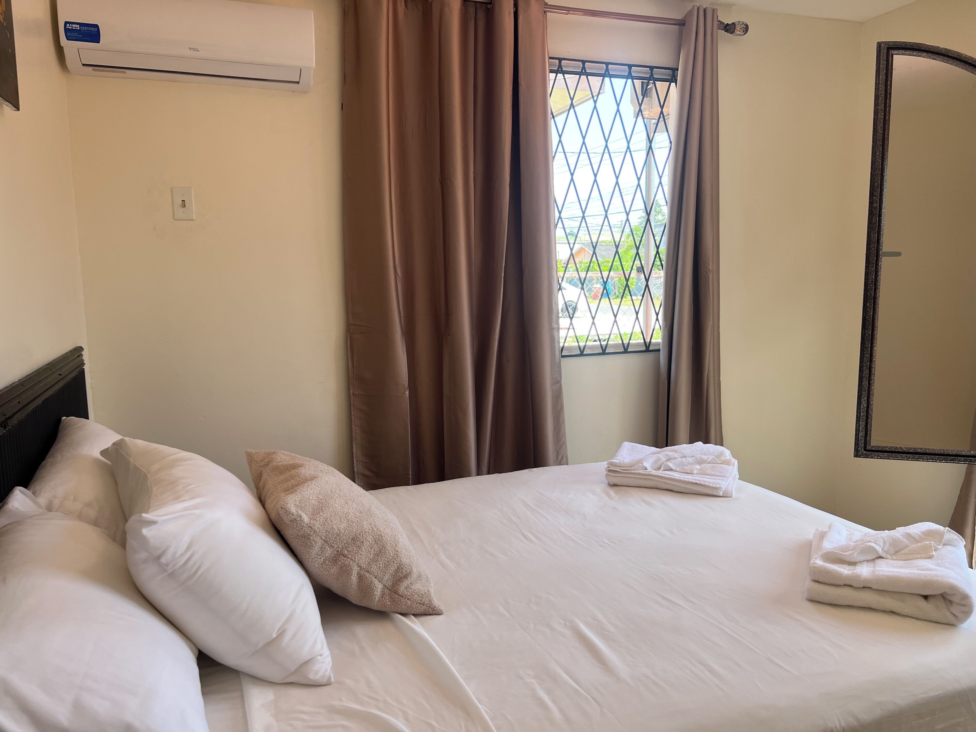 Une chambre d’hôtel avec un lit, des oreillers blancs, une serviette pliée, un appareil de climatisation, une fenêtre avec des rideaux et une porte.