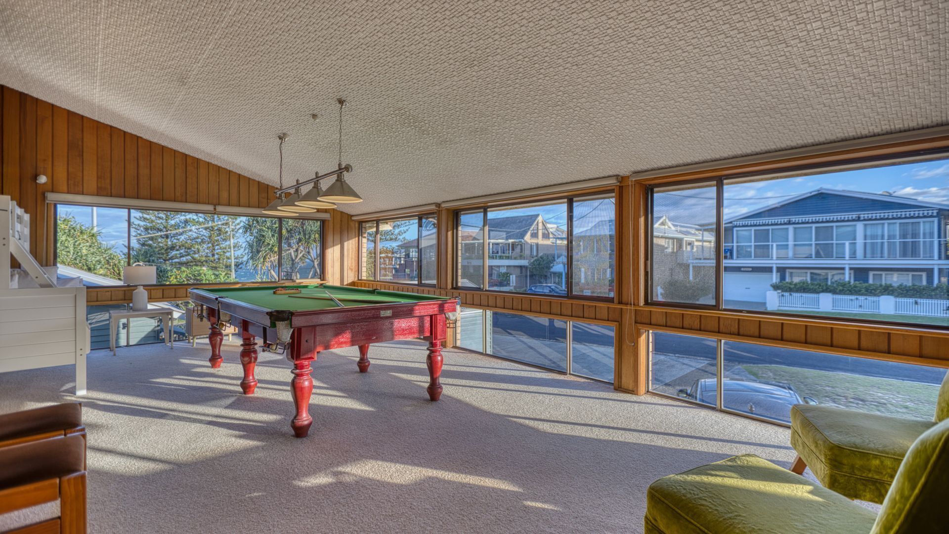 A pool table in a room with large windows, wooden walls, and a view of houses outside.