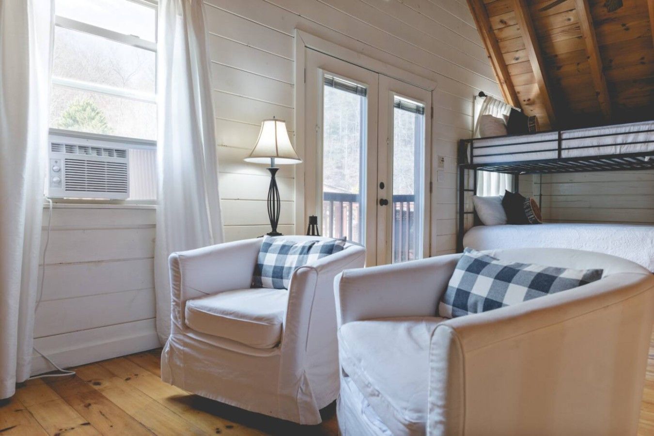 Una habitación acogedora con dos sillones blancos, una lámpara, una cama litera y una ventana con cortinas.