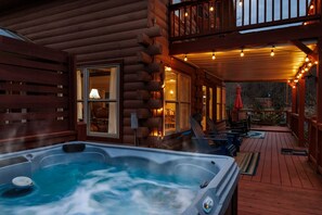 A log cabin with a hot tub on the deck, wooden furniture, and string lights.