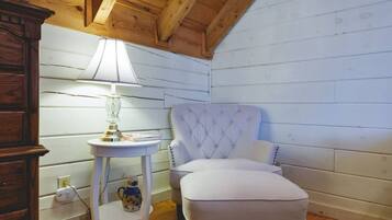 A cozy room with a wooden ceiling, a white upholstered chair, a small round table with a lamp, and a decorative pitcher.