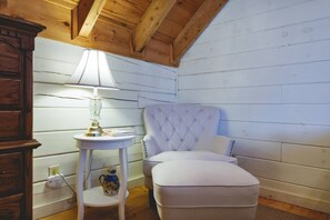 A cozy room with a wooden ceiling, a white upholstered chair, a small round table with a lamp, and a decorative pitcher. - kingisher river cabin (Murphy)
