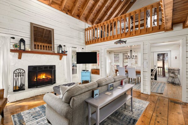 A cozy living room with a fireplace, a television, and a loft area with a wooden railing.