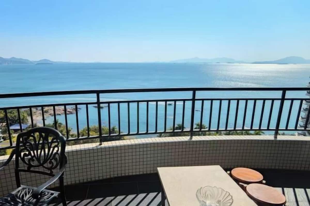 A balcony with a view of the sea, a tiled floor, a table with a decorative bowl, and two stools.