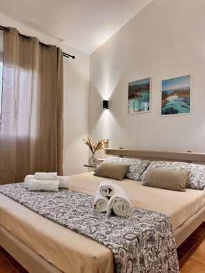 Een netjes opgemaakt bed met patroongedekte lakens, een nachtkastje met een vaas gedroogde bloemen en twee ingelijste foto’s aan de muur.