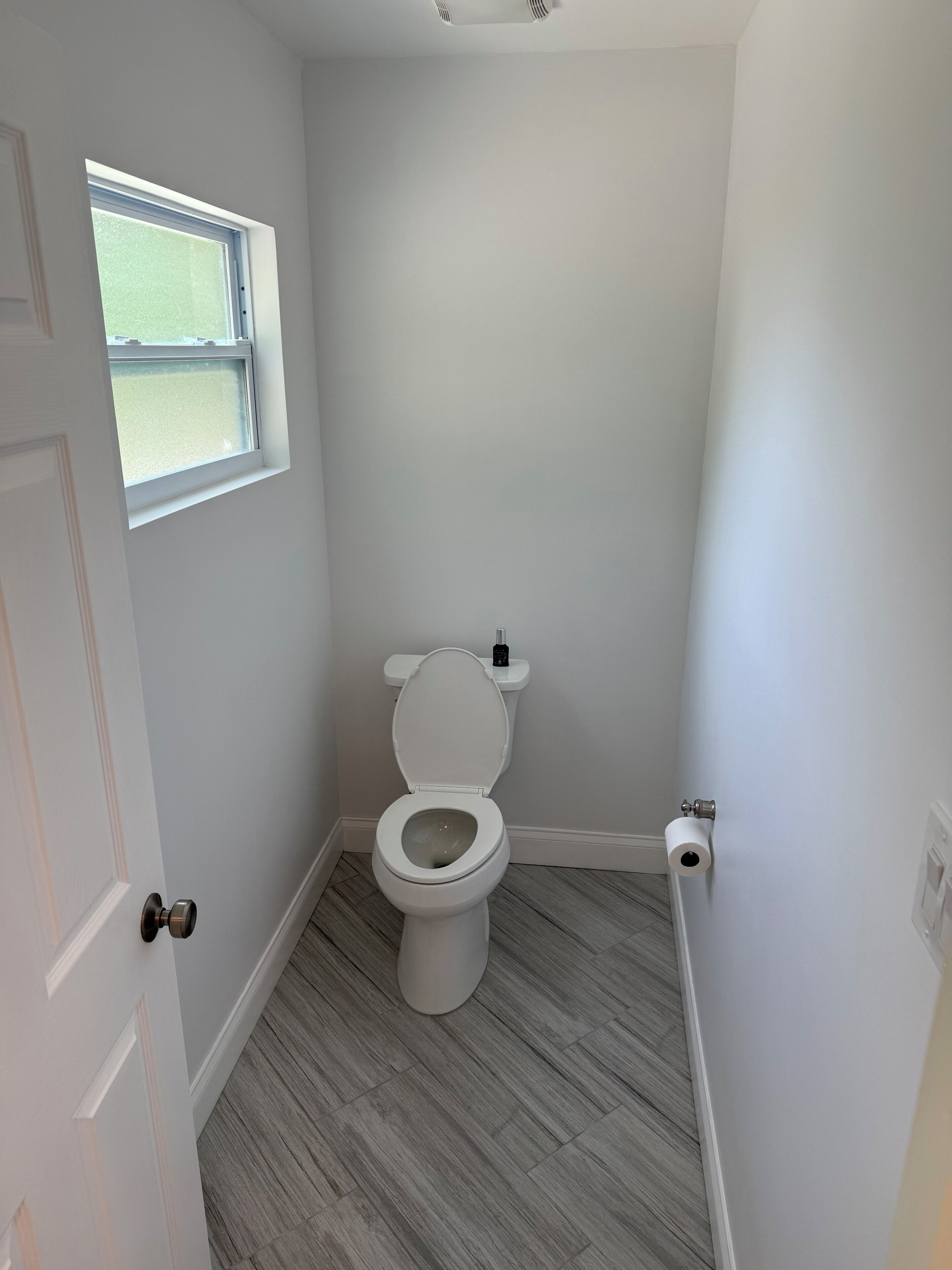 A small, white bathroom with a toilet, a window, and a door.
