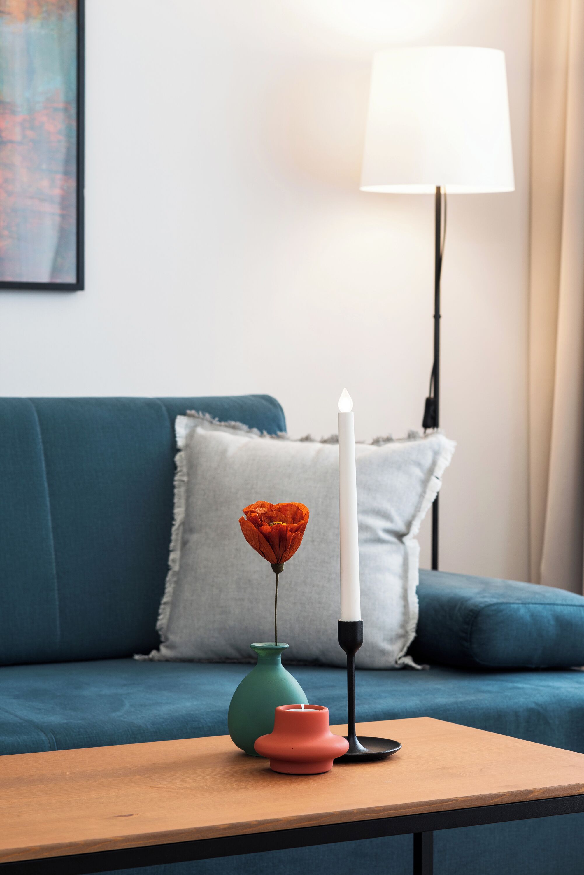 A blue sofa with a grey pillow, a white candle, and a small vase with a flower on a wooden table.