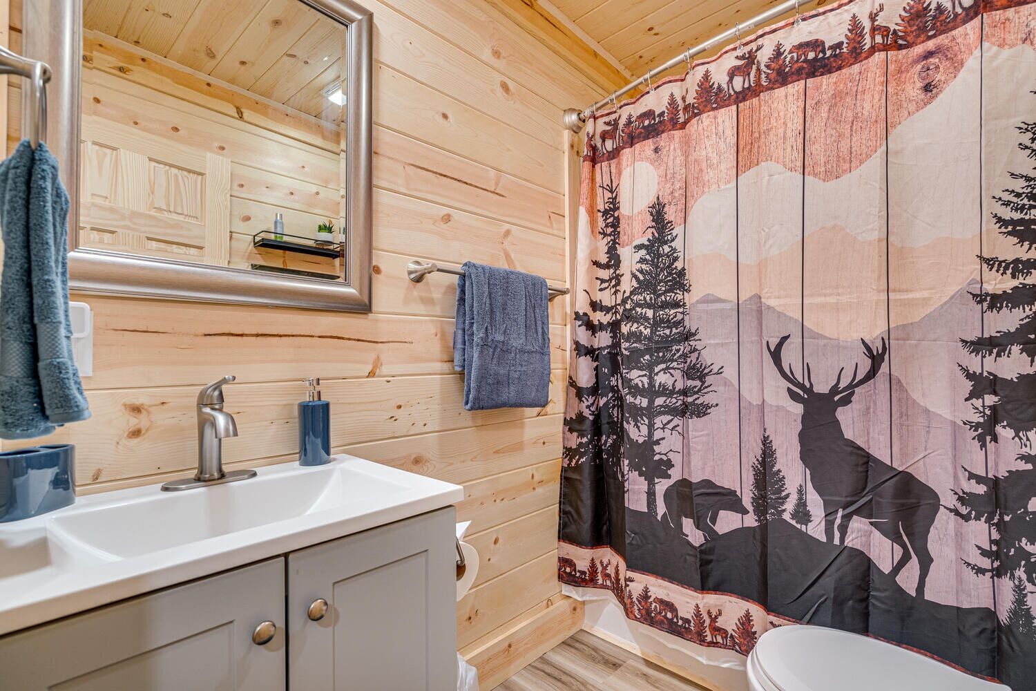A wooden cabin bathroom with a sink, mirror, and shower curtain featuring a deer and forest design.