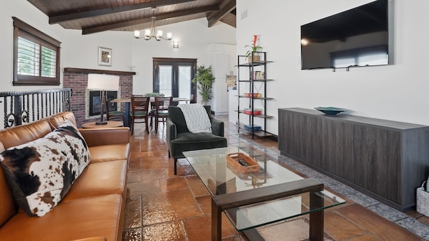 A living room with a leather sofa, a glass coffee table, a fireplace, a TV mounted on the wall, and a bookshelf. - NEW! 3 Houses 2 the Sand-Spacious Gorgeous Remodel (Oxnard)