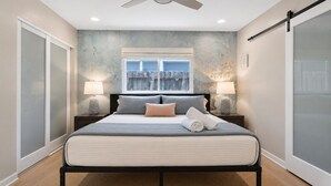 Un dormitorio con una cama, dos lámparas de noche, una ventana con vistas, un ventilador de techo y una pared con textura.