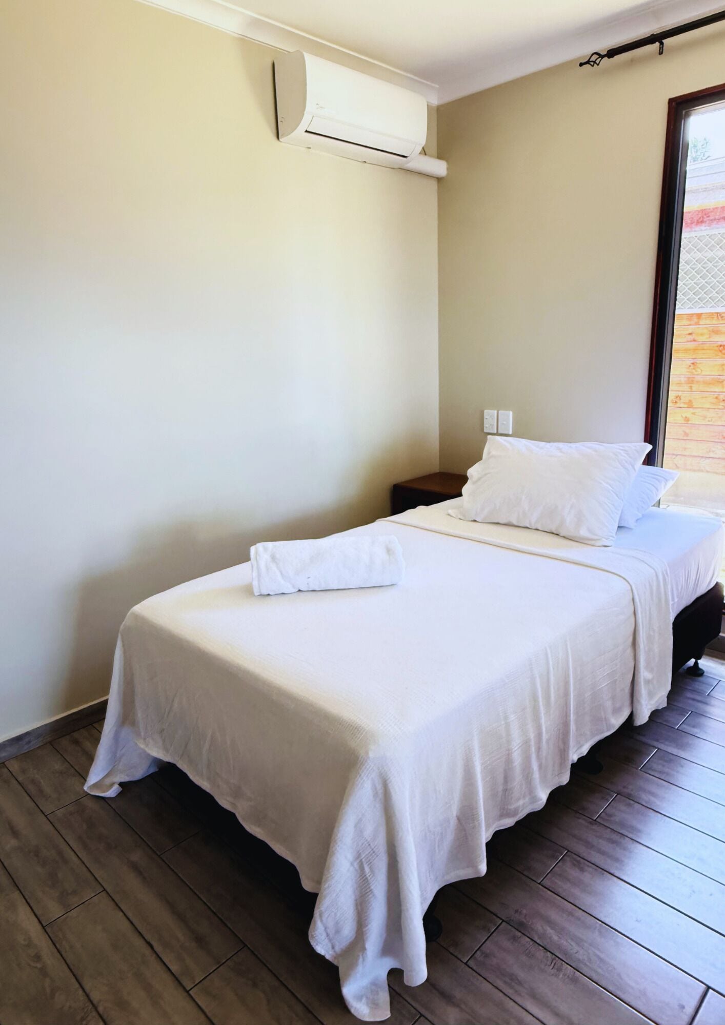 A single bed with white bedding and a white towel, a small bedside table, and a wall-mounted air conditioning unit.