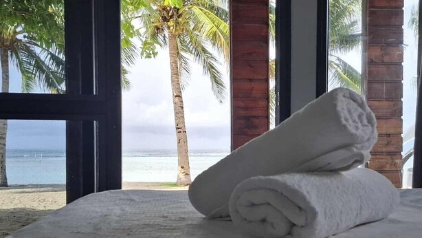 A bed with rolled white towels and a view of the beach and palm trees through large windows. - Miti Maninoa Ocean Club (Maninoa)