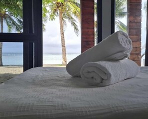 A bed with rolled white towels and a view of the beach and palm trees through large windows.