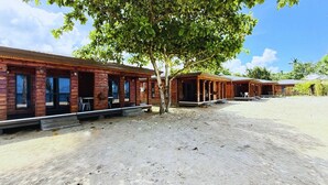 A row of wooden cabins with outdoor seating, a sandy ground, and a tree in the center. - Miti Maninoa Ocean Club (Maninoa)