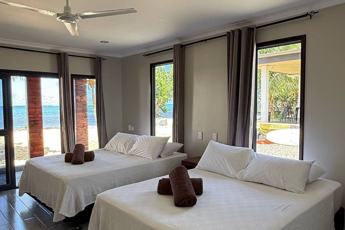 A hotel room with two beds, a ceiling fan, and large windows offering a view of the beach.