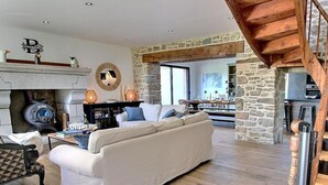 A spacious living room with a stone fireplace, a sofa, and a wooden staircase. - La Marie Morgane - LEZARDRIEUX (LEZARDRIEUX)