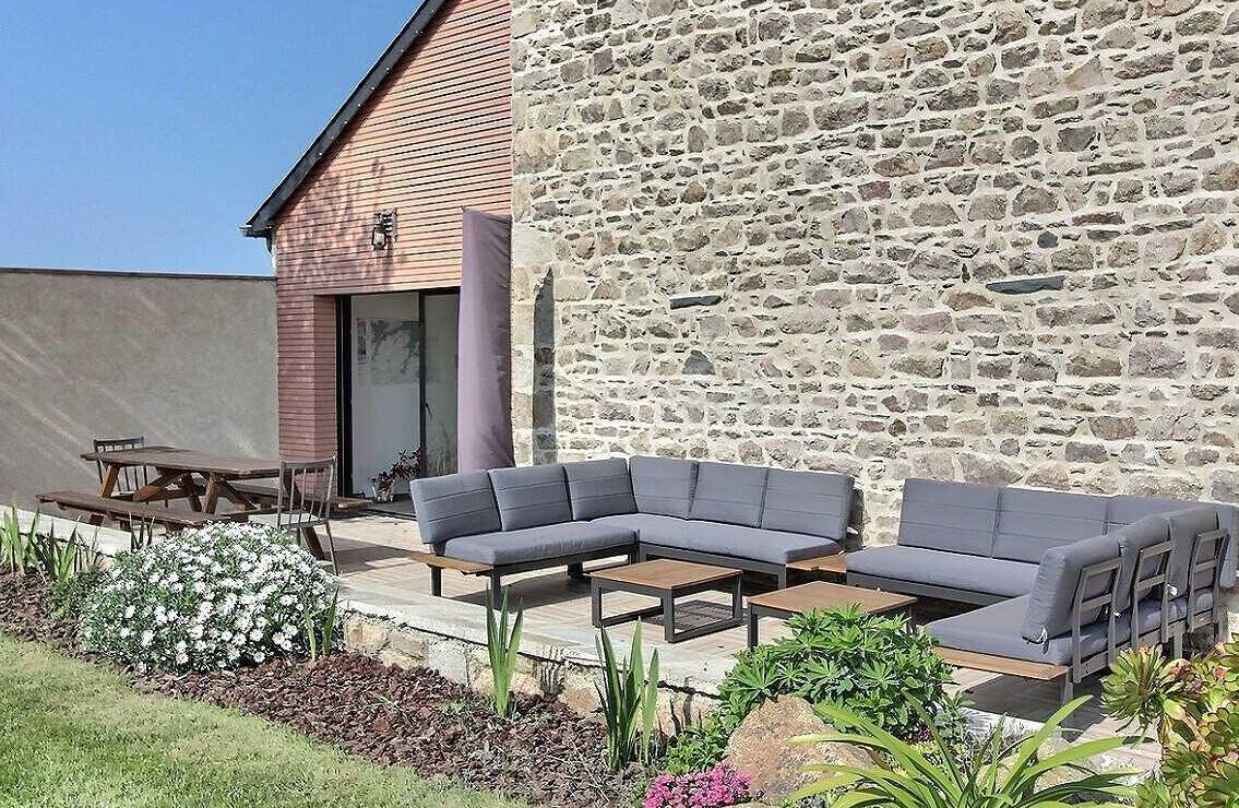 A stone wall with a wooden building adjacent to it, a grey sectional sofa, and a wooden table and chairs in a garden setting.