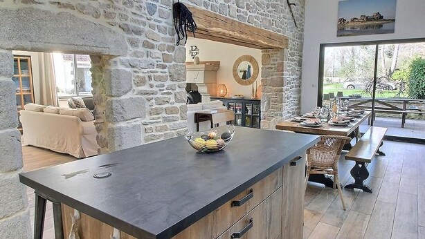 A stone-walled kitchen with a central island, a dining area with benches, and a view of the outdoors. - La Marie Morgane - LEZARDRIEUX (LEZARDRIEUX)