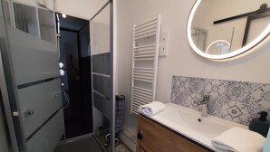 A modern bathroom with a glass shower enclosure, a white vanity with a patterned tile backsplash, and a round mirror with a light.