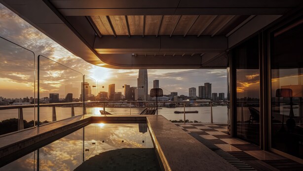 Un bâtiment moderne à la façade vitrée, doté d’une piscine et offrant une vue sur les toits de la ville au coucher du soleil.