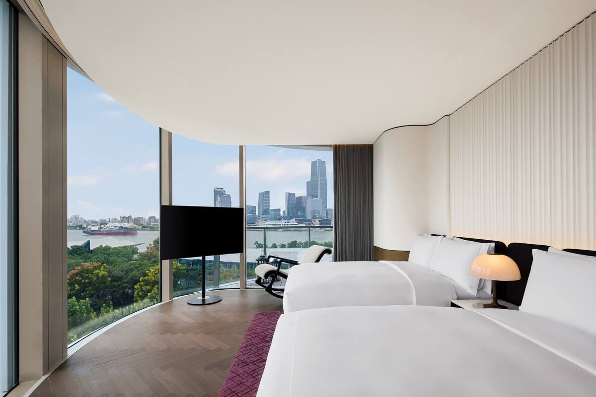 Ein modernes Hotelzimmer mit großem Fenster und Blick auf die Stadt, einem Flachbildfernseher, einem bequemen Bett und einem Schreibtisch mit Stuhl.