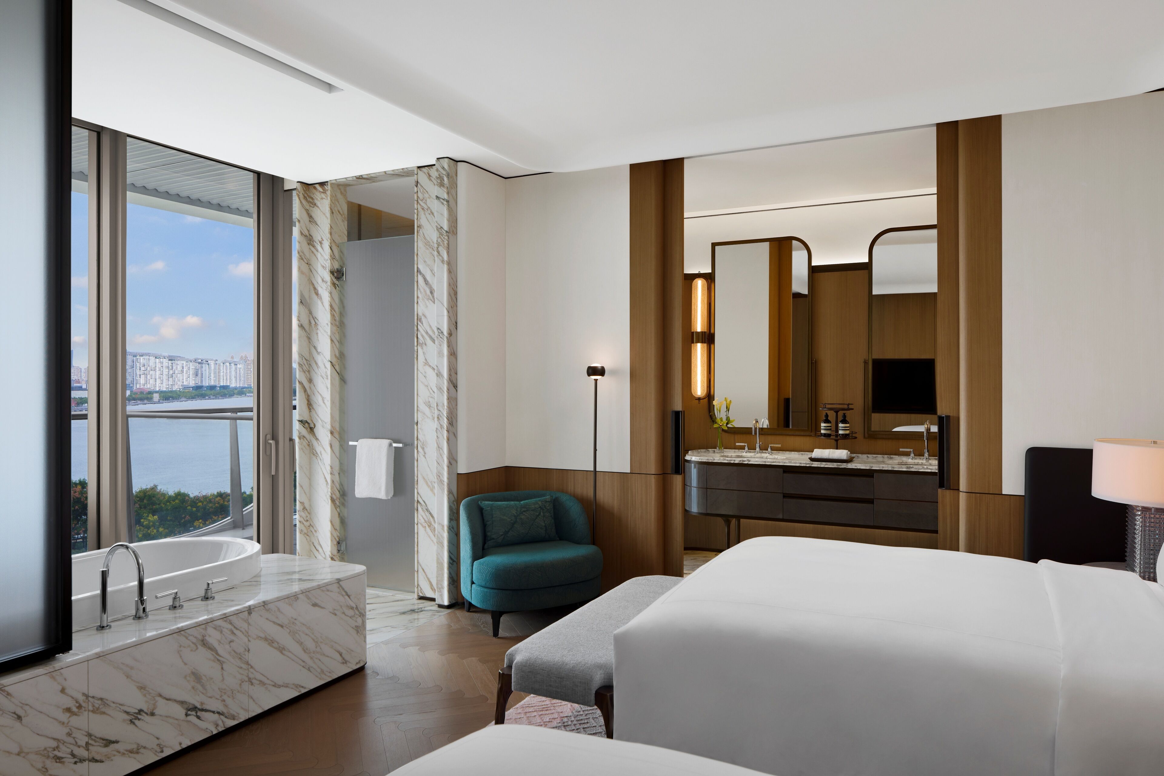A modern hotel room with a large window, a marble bathroom, a bed, a chair, and a vanity with a mirror.