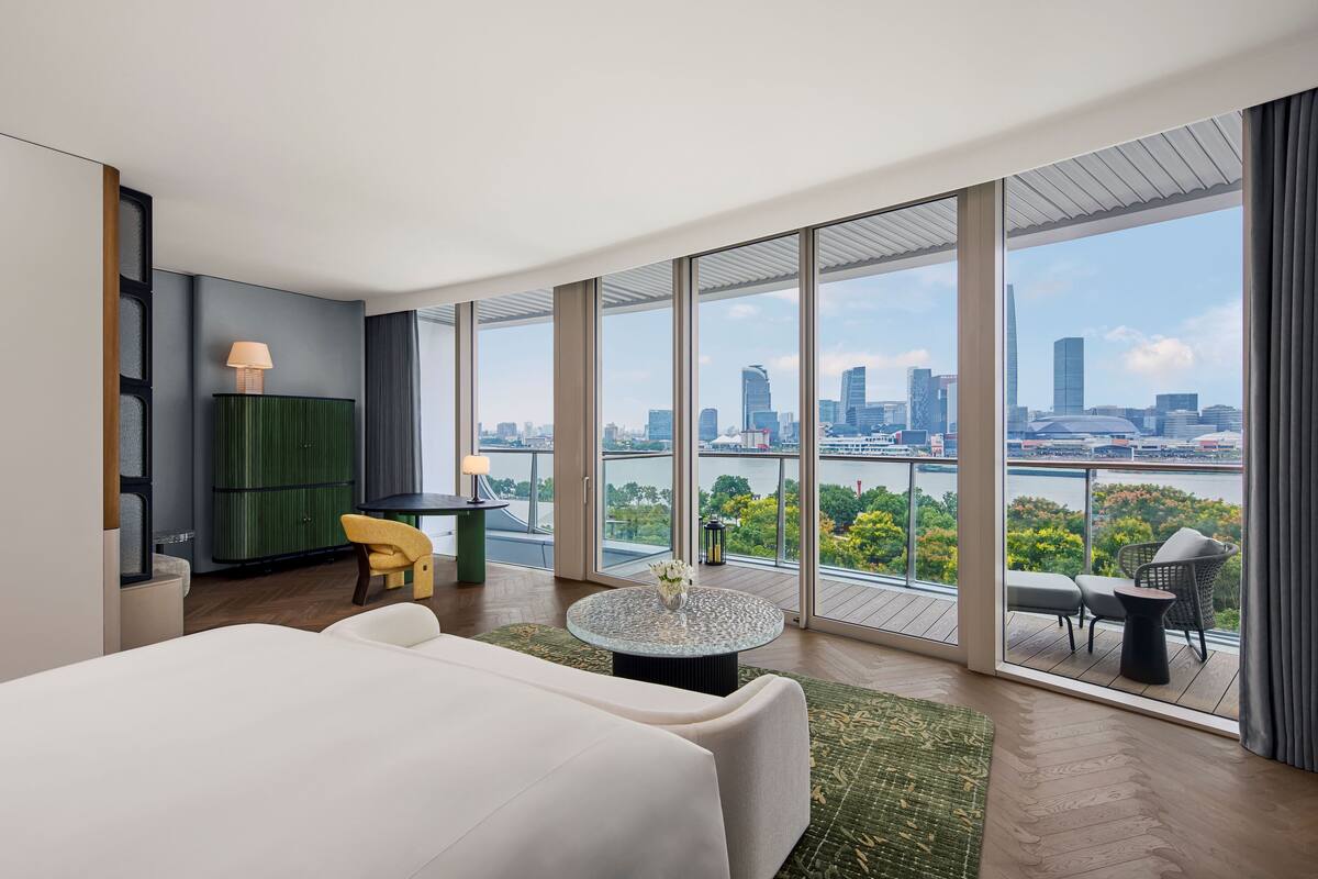 Ein modernes Hotelzimmer mit einem großen Bett, einem runden Tisch mit einer Vase und Blick auf eine Skyline und einen Fluss.