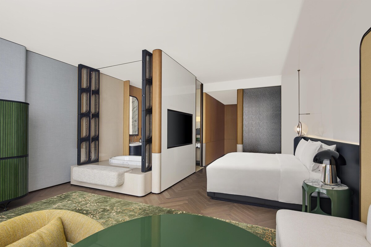 A modern hotel room with a bed, a television, a bathroom with a sink, and a green accent wall.