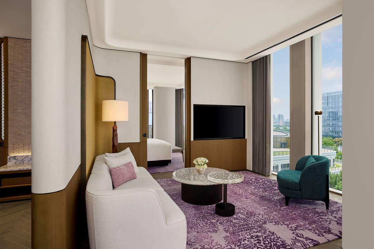 Ein modernes Hotelzimmer mit einem weißen Sofa, einem runden Couchtisch, einem türkisblauen Sessel, einem großen Fernseher und Blick auf die Stadt durch die Fenster.