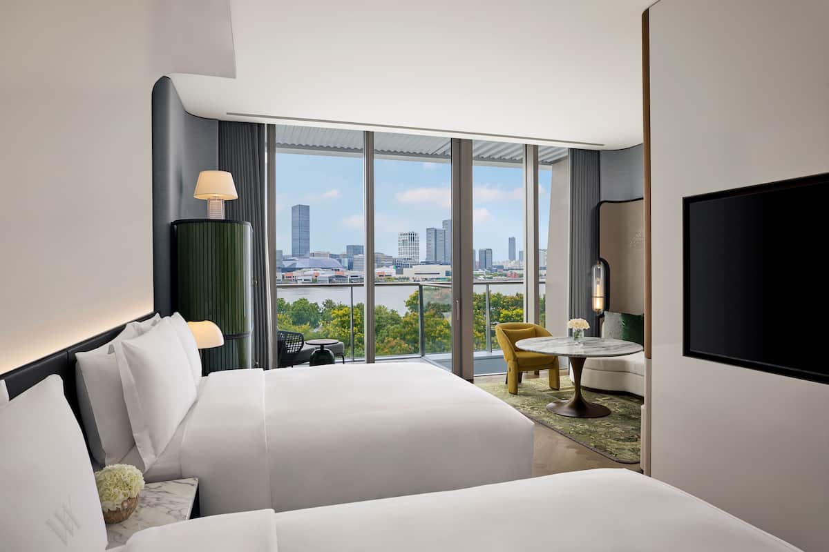 Ein modernes Hotelzimmer mit einem großen Bett, einem kleinen Tisch, einem Sessel und einem Fernseher. Das Zimmer hat ein großes Fenster mit Blick auf die Stadt.