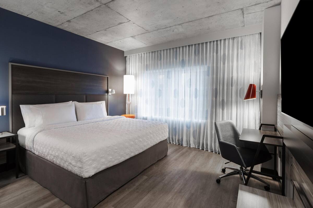 Habitación de hotel con una cama grande, un escritorio, una silla, una mesita de noche y un televisor.