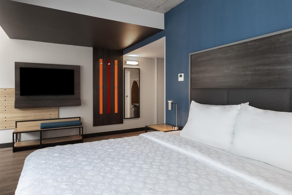 Una habitación de hotel moderna con una cama grande, un televisor de pantalla plana, un banco de madera y una obra de arte única en la pared.
