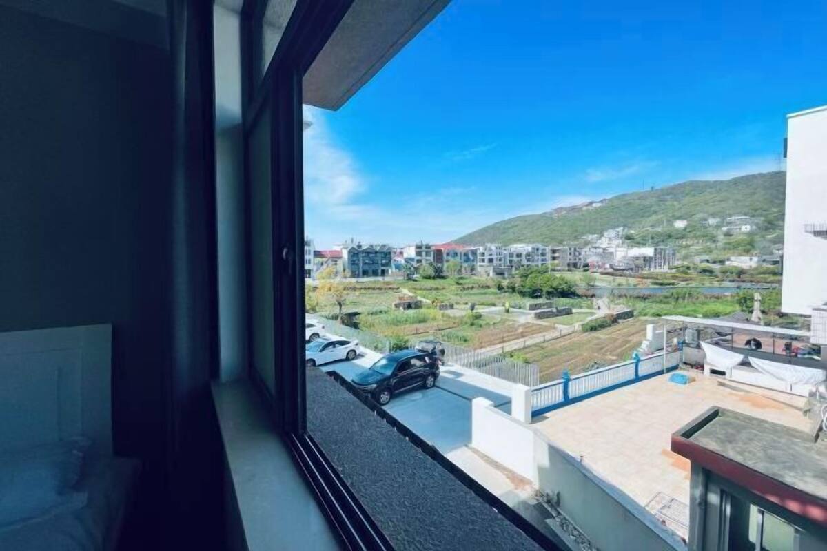 Blick aus einem Fenster auf einen Parkplatz mit Autos, ein Gebäude mit weißer Fassade und eine grüne Landschaft mit Hügeln im Hintergrund.