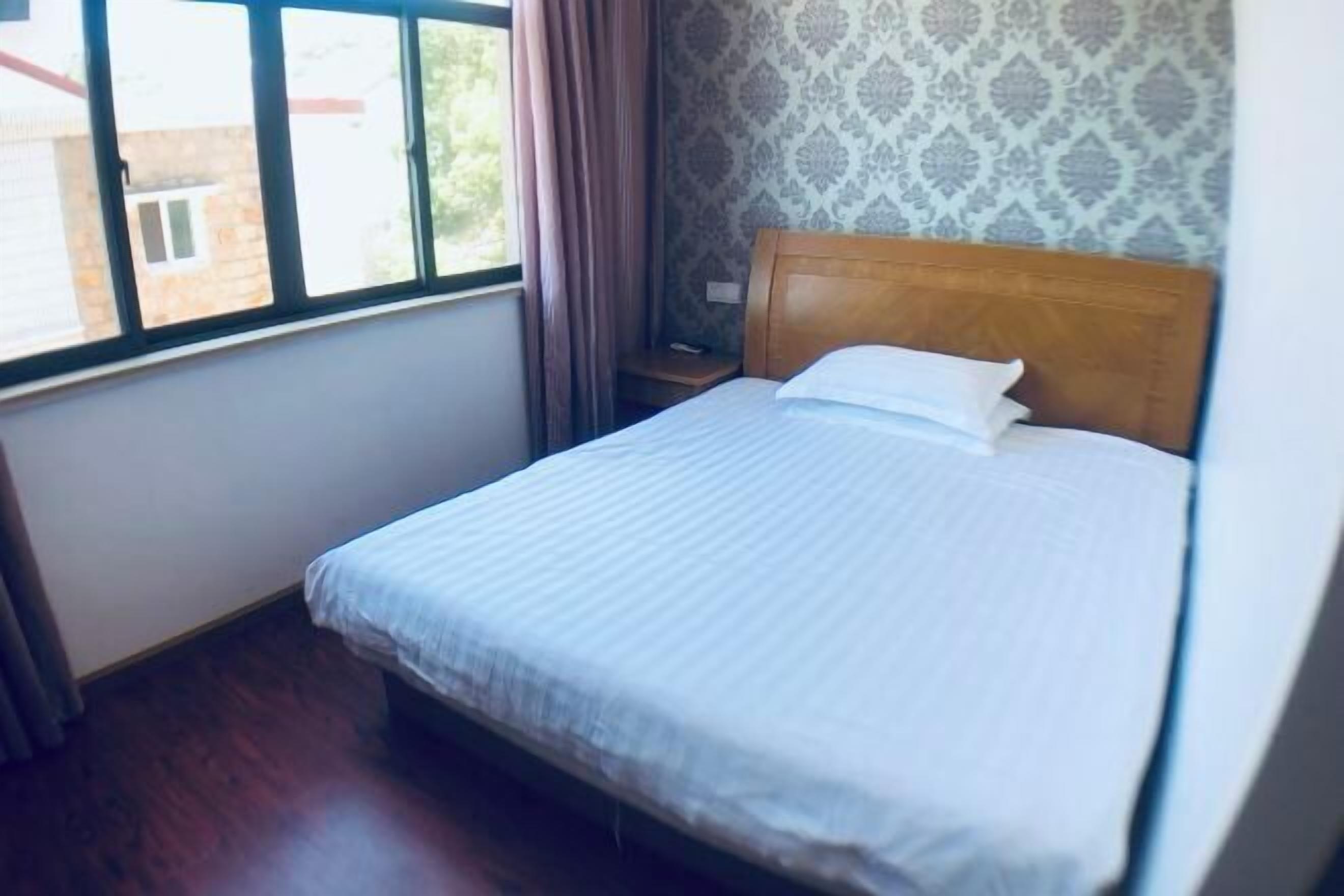 A single bed with a wooden headboard, a nightstand, a window with curtains, and patterned wallpaper.