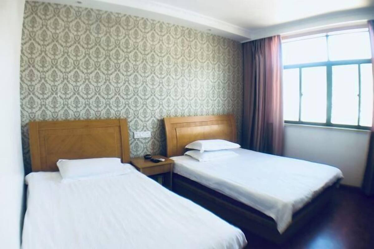 Ein Hotelzimmer mit zwei Betten, einem Wandteppich mit Muster, einem Fenster mit Vorhängen und einem Nachttisch mit Telefon.